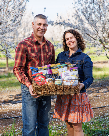 Two people holding a basket full of nuts and nut butters in an almond orchard with cherry blossom trees.