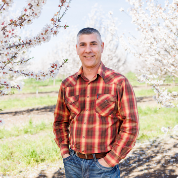 Isidro in a plaid shirt standing in an orchard with blooming trees